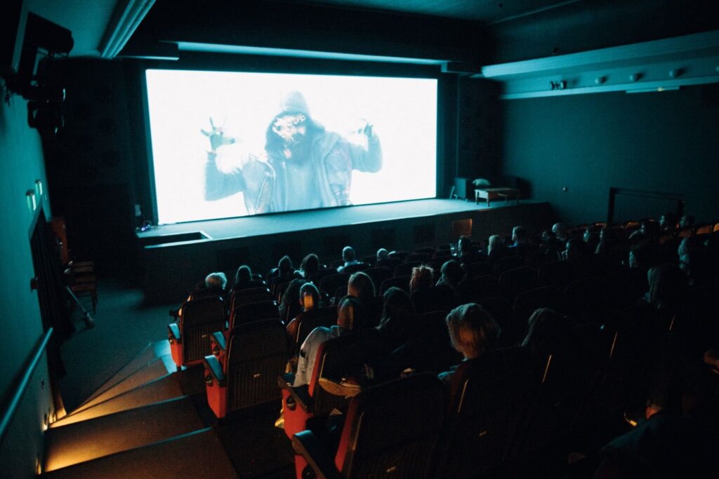 Audience in a dim auditorium facing a bright screen, stair lights glowing