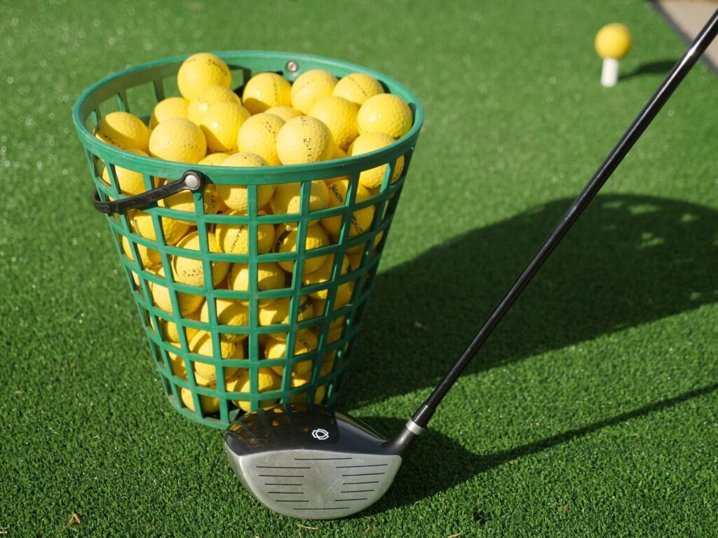 Basket of yellow range balls beside a driver on artificial turf