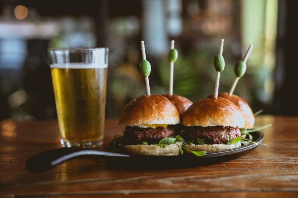 Beef sliders on skewers beside a glass of lager on a wooden table