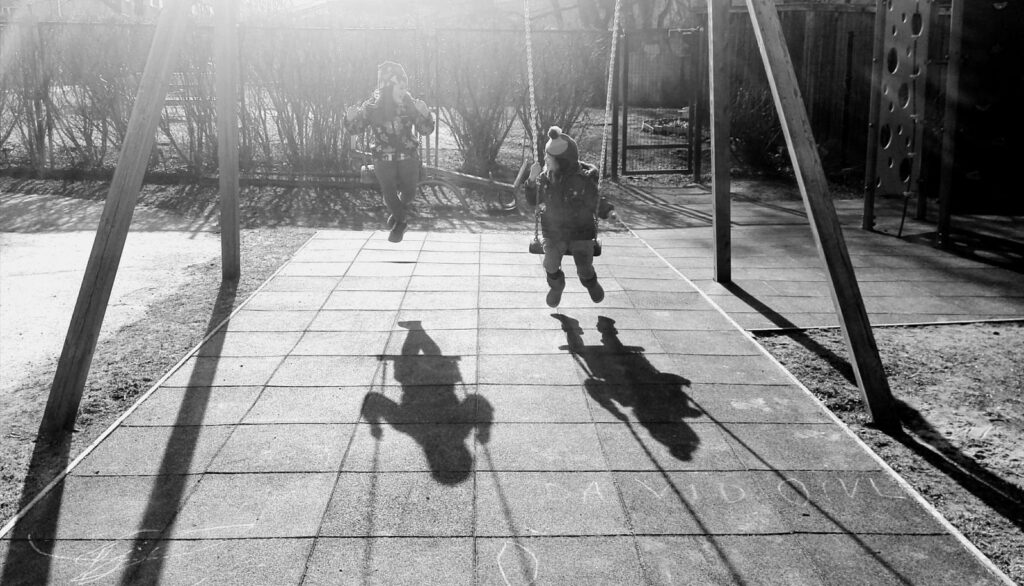 Black-and-white photo of two children on swings with long shadows across the ground