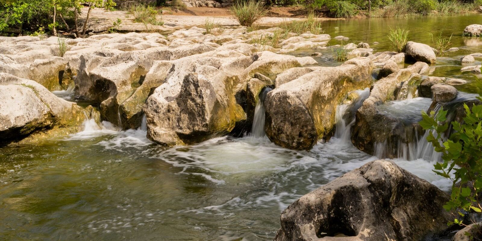 Clear creek water spilling over pale limestone boulders and ledges under green trees