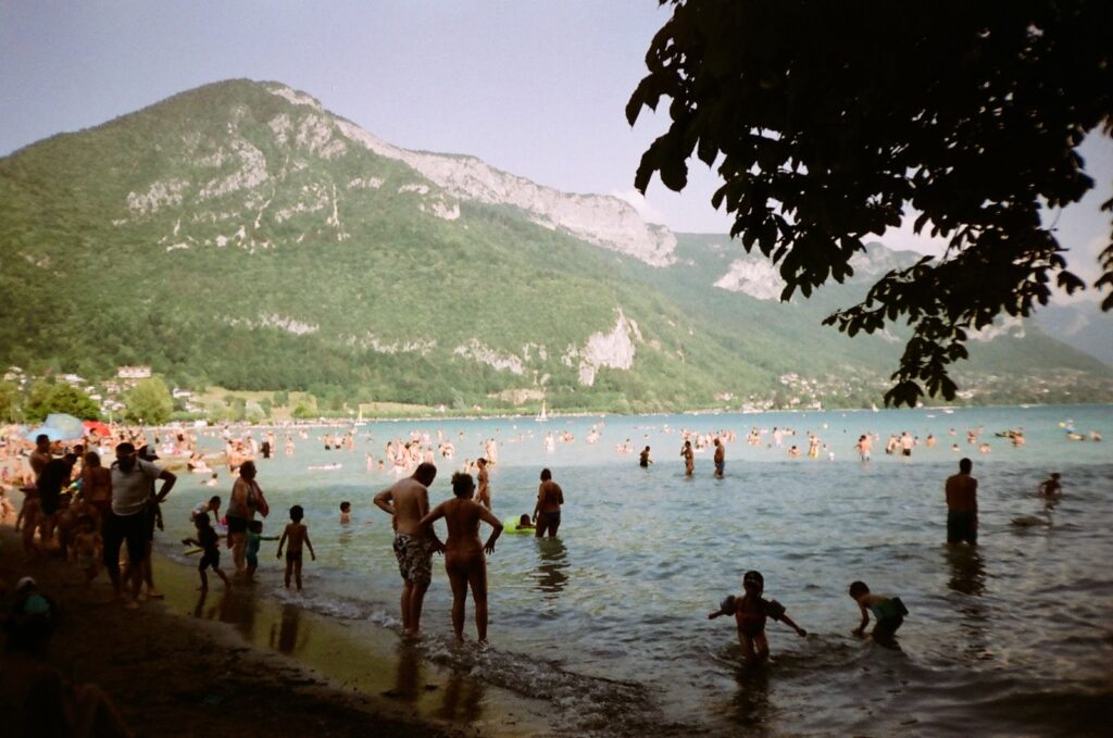 Crowded summer lake beach with swimmers wading in shallow water below a hillside