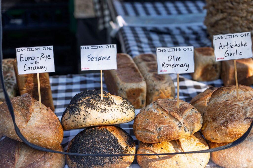Crusty loaves labeled rye, sesame, olive rosemary and artichoke garlic at a bread stall