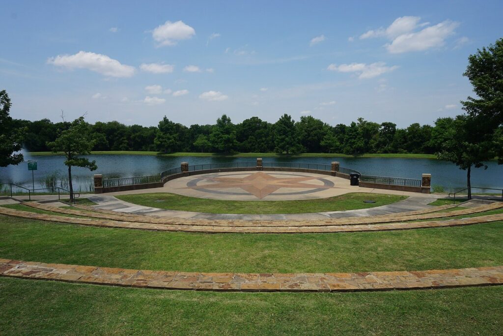 Curved stone amphitheater seating facing the pond at Frisco Commons Park under a blue sky
