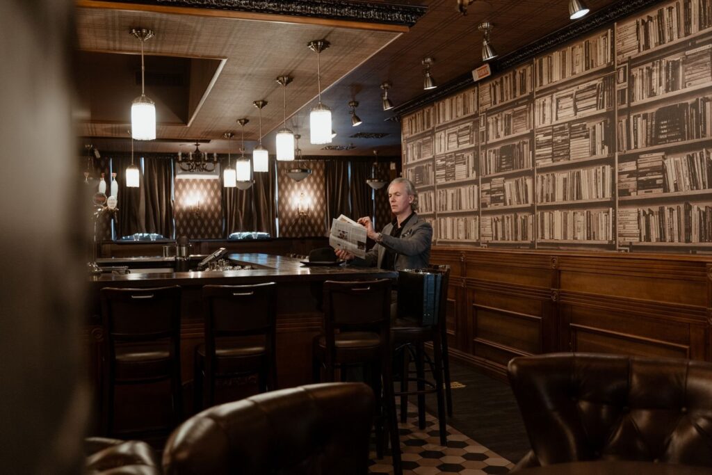 Dim bar room with leather stools, pendant lamps and bookshelf-print wallpaper