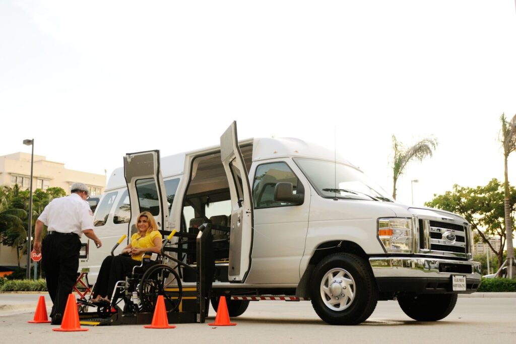 Driver helping a wheelchair user board a white van with a hydraulic lift