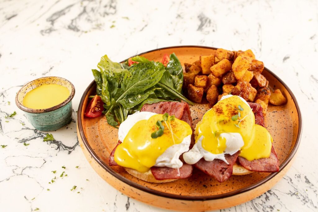 Eggs benedict with hollandaise, ham, roasted potatoes and greens on an orange plate