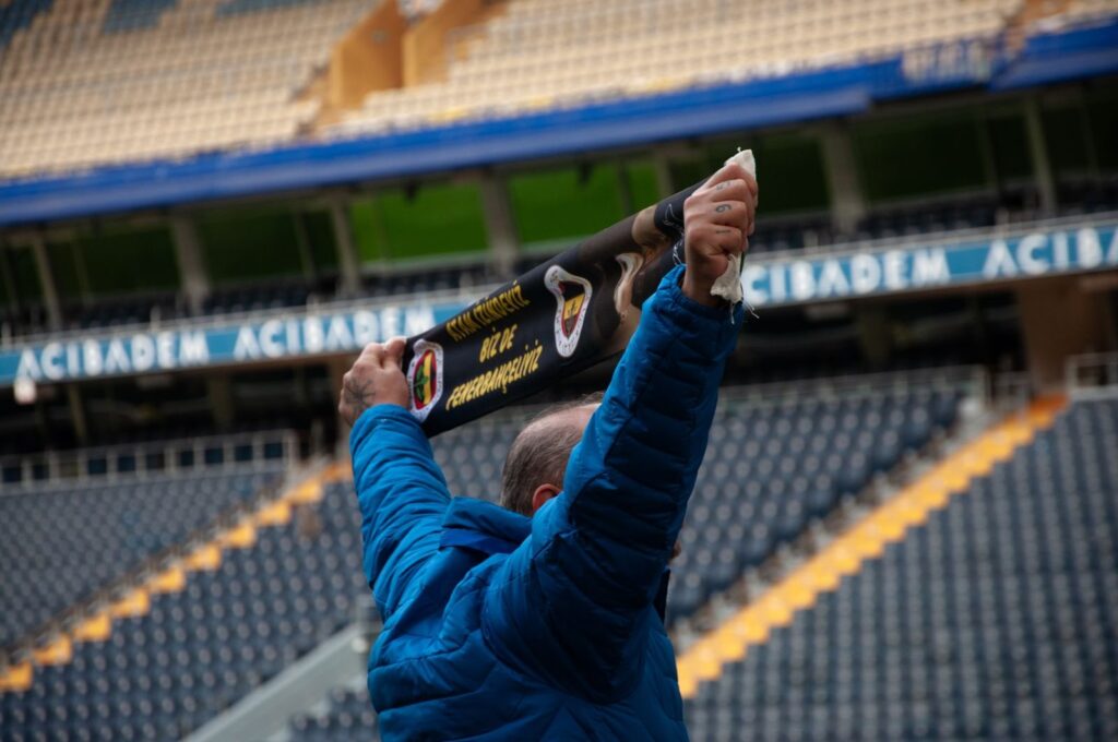 Fan in a blue jacket holding a supporters' scarf overhead in an empty stadium
