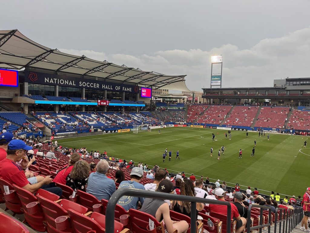 Fans in red seats watching a match at Toyota Stadium beneath the National Soccer Hall of Fame sign
