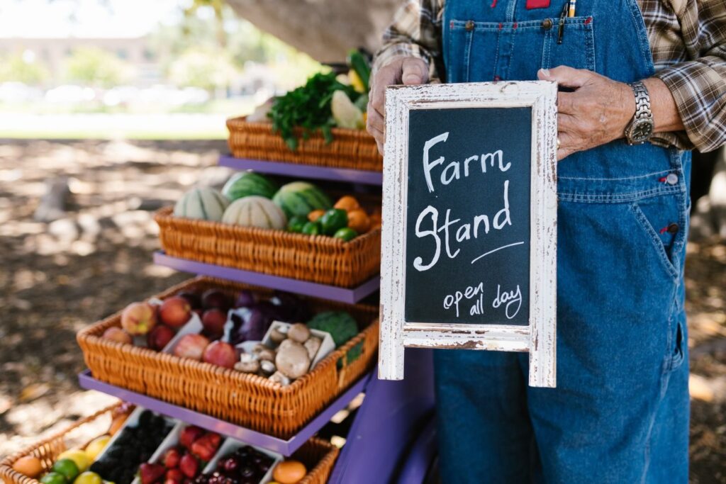 Farmer holding a chalkboard farm stand sign beside baskets of fresh fruit and vegetables