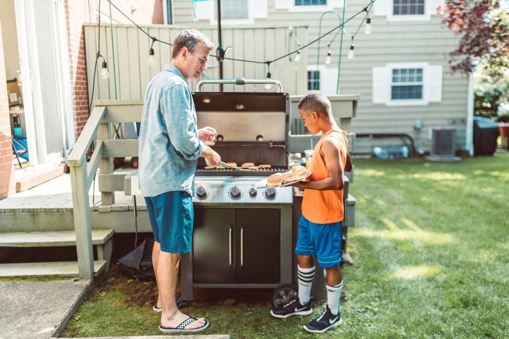 Father and son grilling on a gas barbecue in a fenced backyard strung with lights
