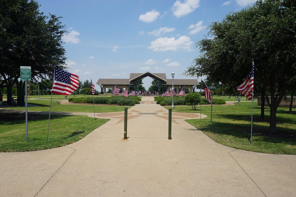 Flag-lined concrete walkway leading to a large arched pavilion in a Frisco park