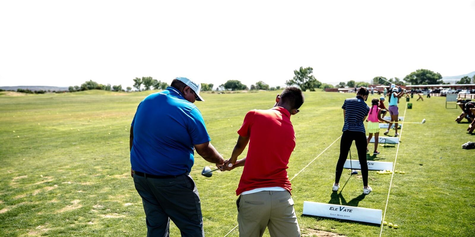 Golf instructor guiding a student's grip on a driving range with a group clinic practicing behind them