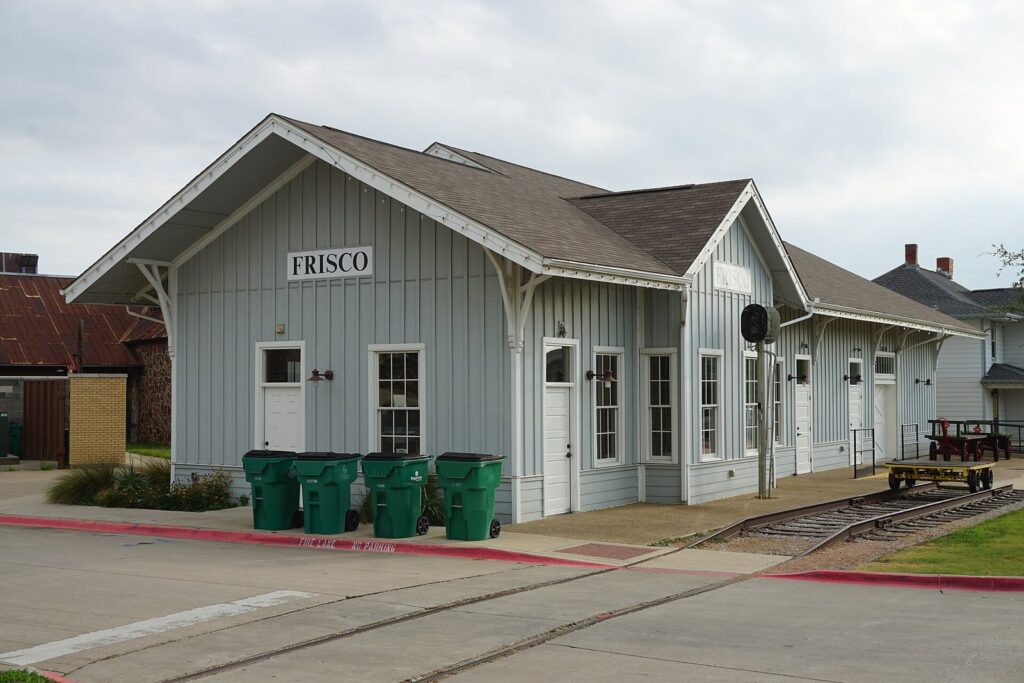 Gray wooden railroad depot with a Frisco sign and a baggage cart on the rails at the Frisco Heritage Center