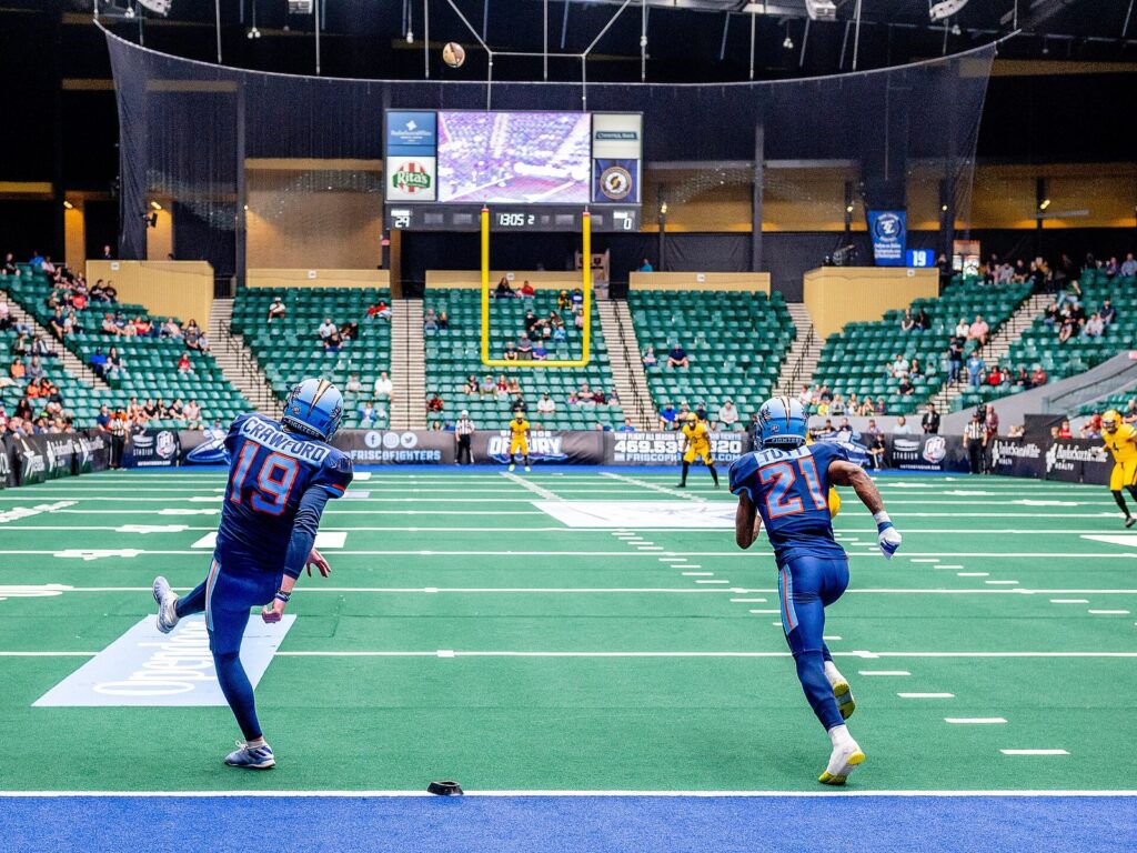Indoor football players in blue running across turf inside the Comerica Center arena