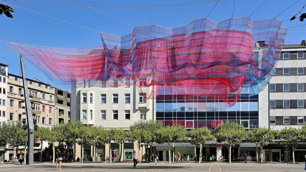 Janet Echelman's red and blue net sculpture suspended above a city square in Frankfurt