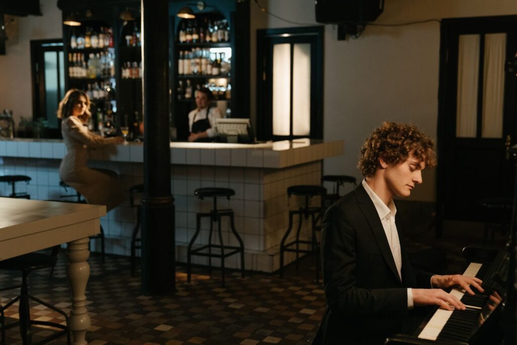 Man in a black suit playing piano inside a bar while a woman sits at the counter