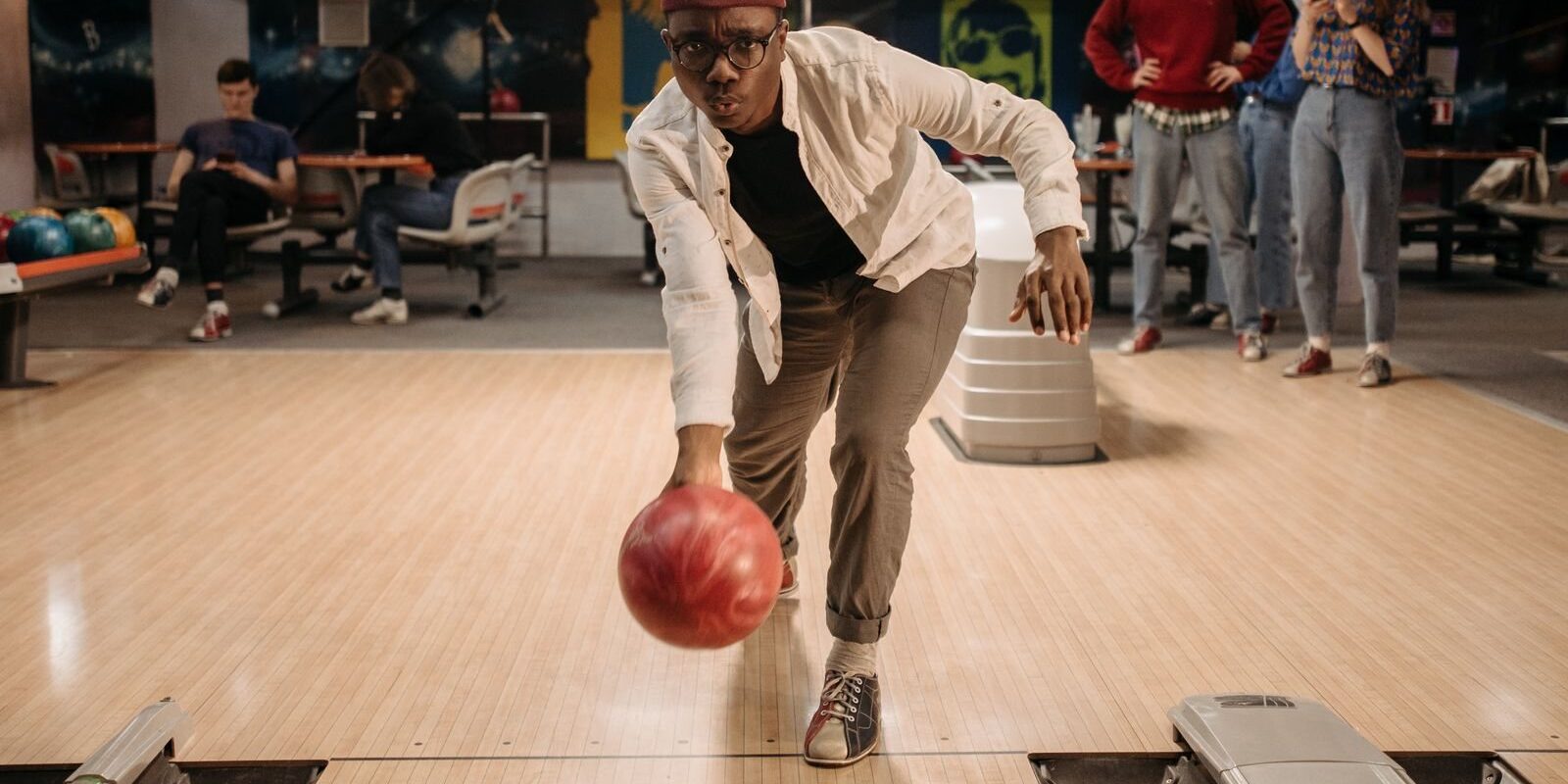 Man rolling a red ball down the lane while friends wait by the ball return