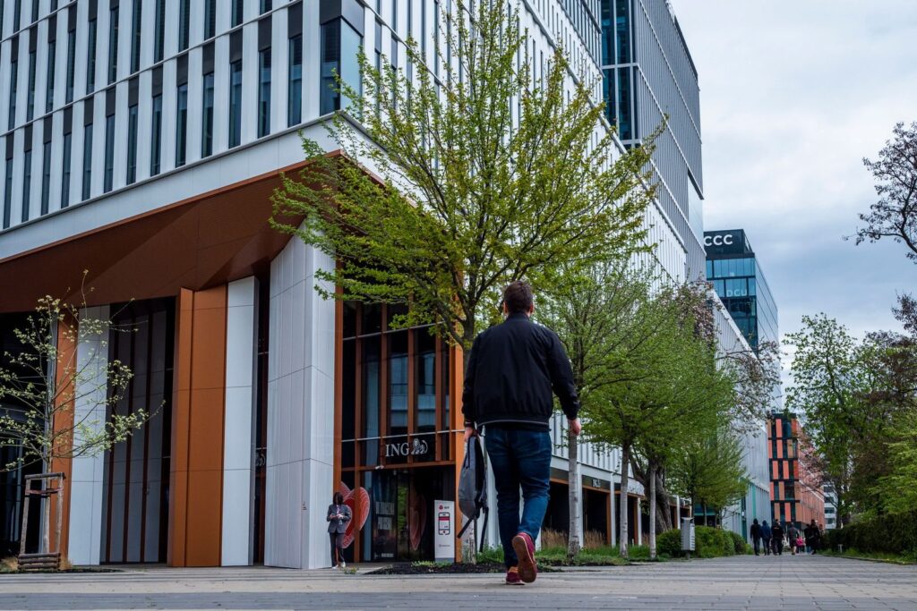 Man walking along a tree-lined sidewalk past modern mid-rise buildings