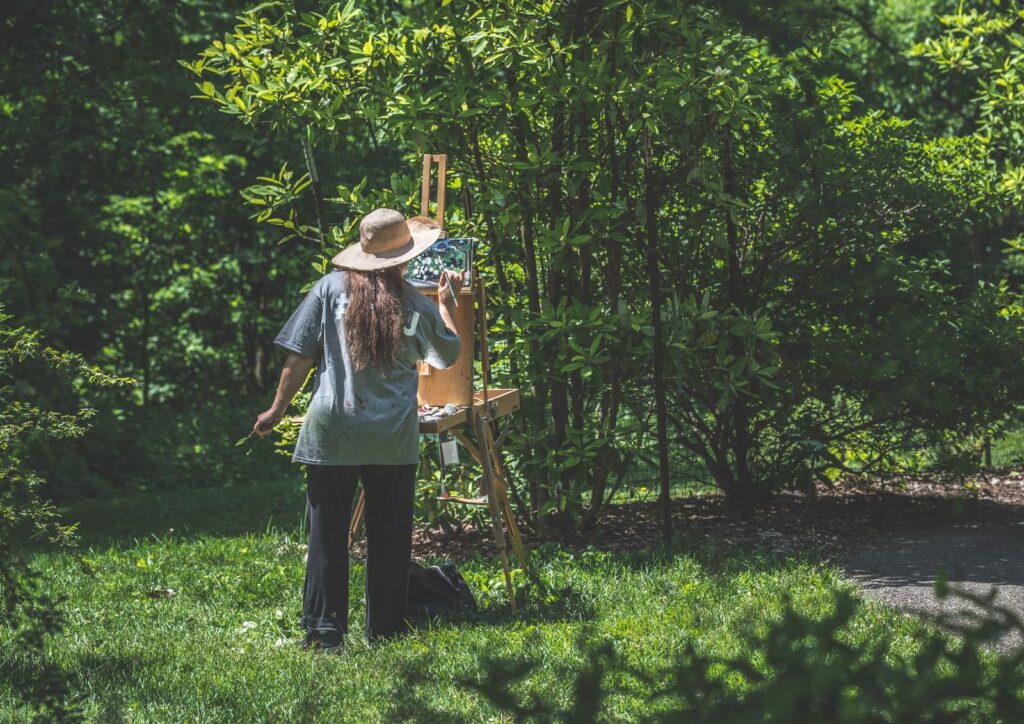 Painter in a sun hat working at an easel on the grass in a leafy park