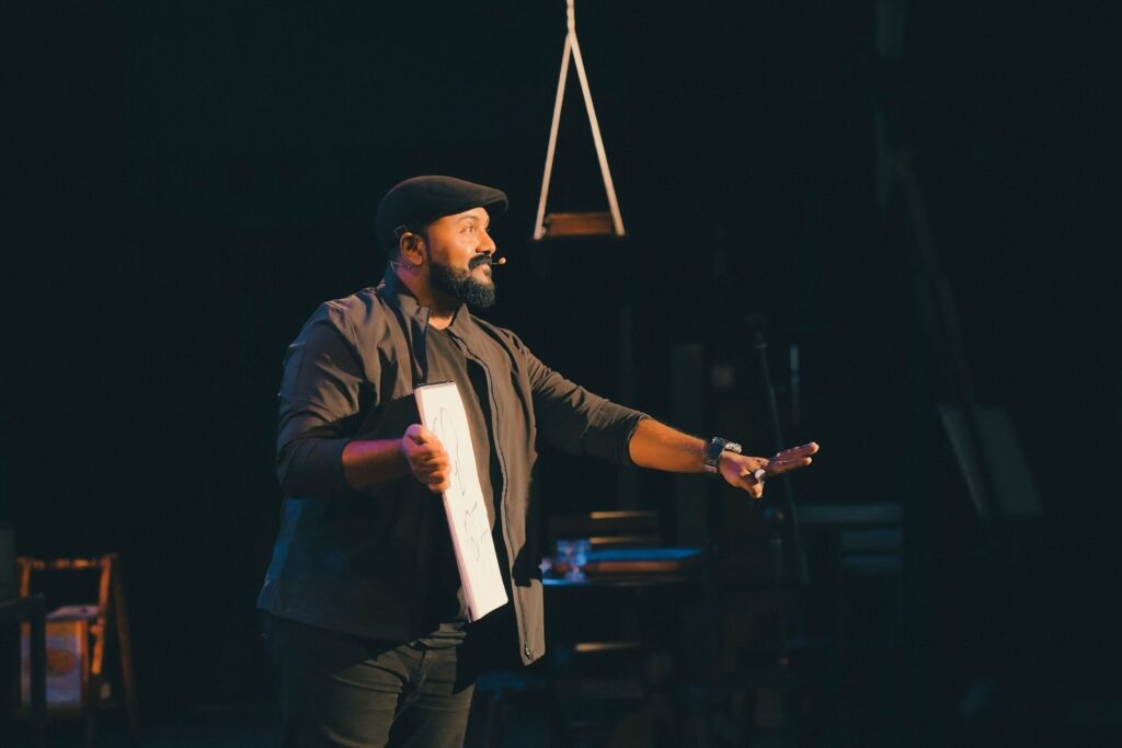 Performer in a flat cap holding a script under a single spotlight on a dark stage