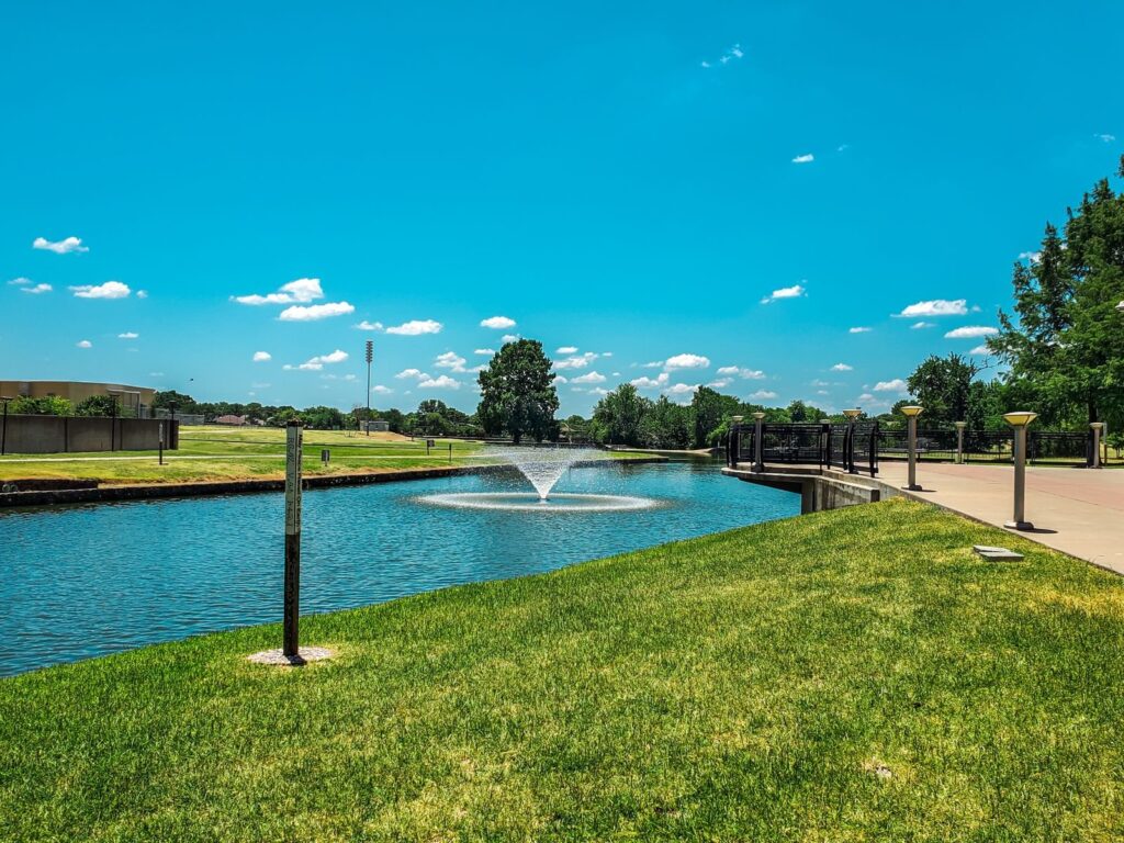 Pond with a spray fountain beside a paved walkway and green lawn under a blue sky