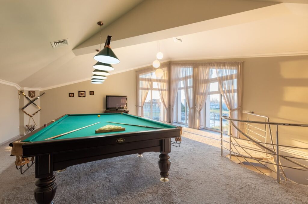 Pool table with cues in a carpeted game room with tall curtained windows
