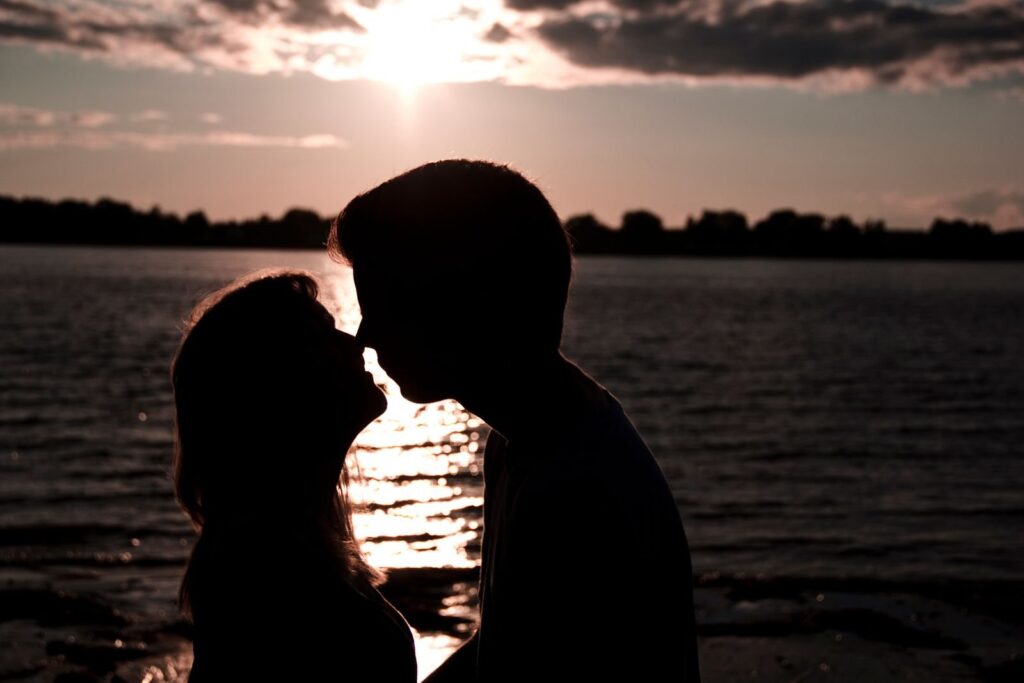 Silhouetted couple leaning in for a kiss beside a lake at sunset