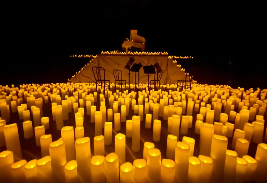 Small stage with a piano and music stands surrounded by hundreds of lit candles