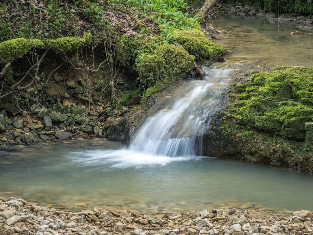 Small waterfall pouring over a mossy rock lip into a shallow gravel-bottomed pool