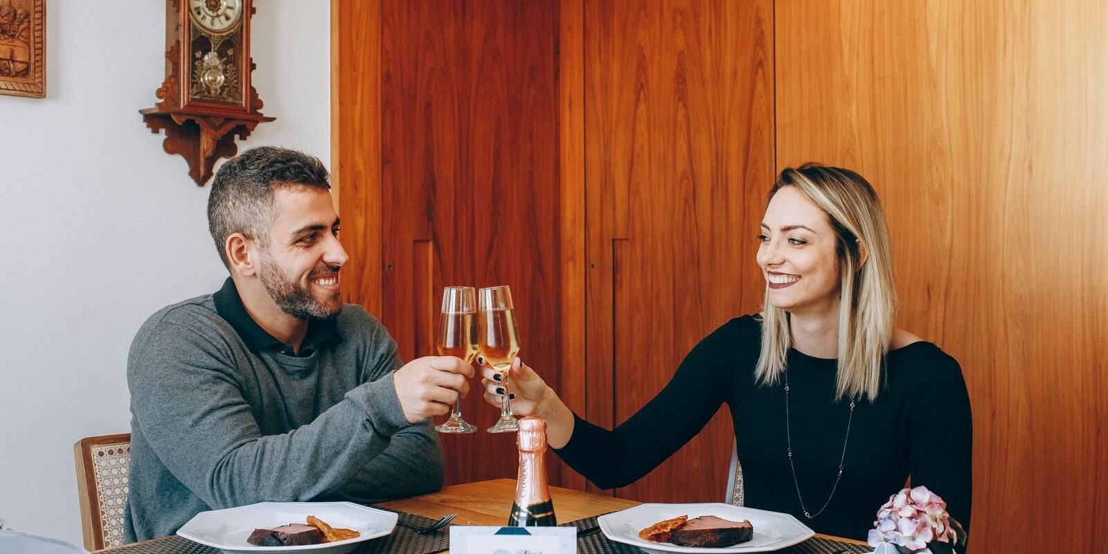 Smiling couple clinking champagne glasses over dinner with a greeting card on the table