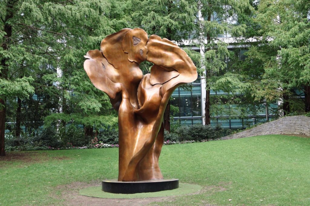 Tall abstract bronze sculpture on a round base in a tree-lined lawn