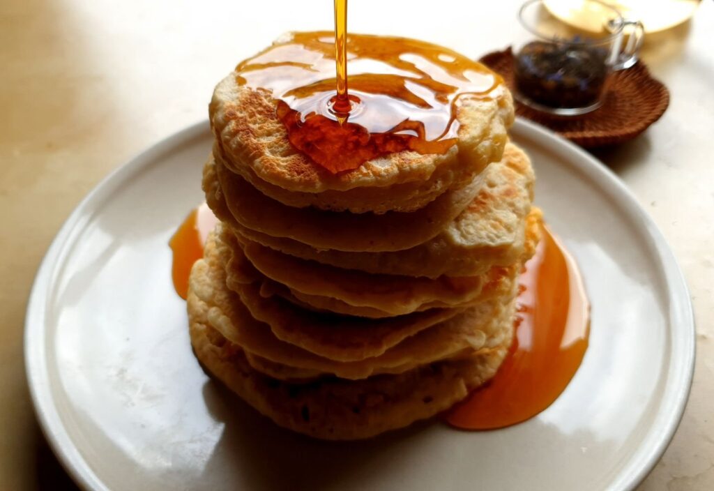 Tall stack of pancakes with syrup pouring over the top on a white plate