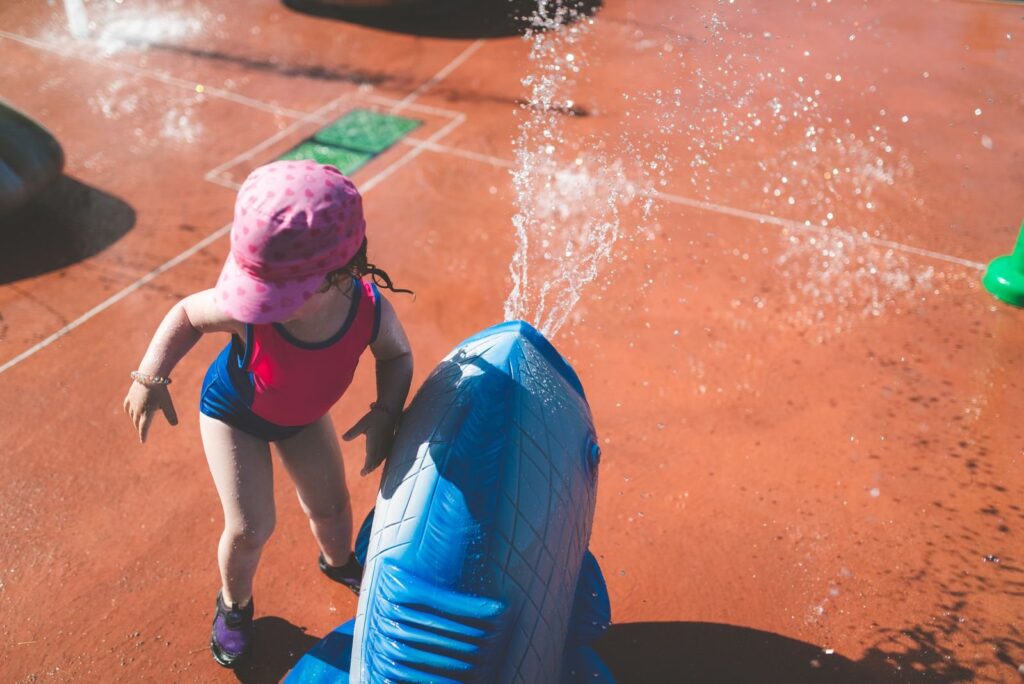 Toddler in a pink sun hat and swimsuit playing beside a spray jet at a splash pad