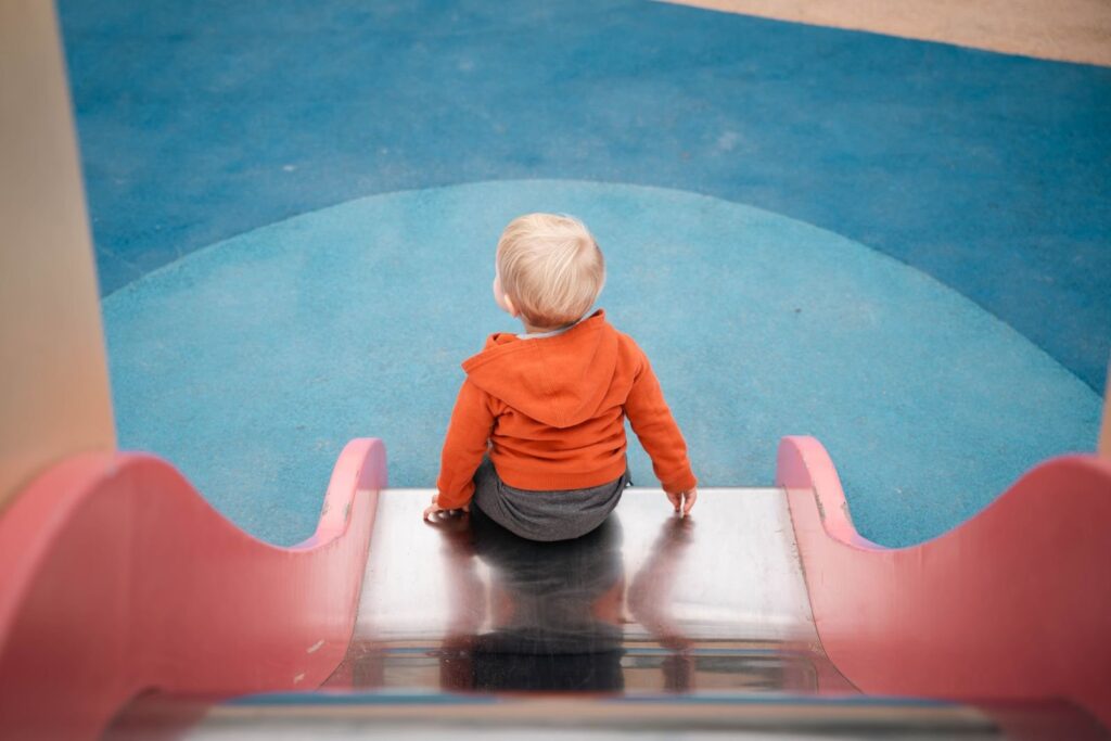 Toddler in an orange hoodie at the top of a pink slide, a staple of Frisco TX toddlers' park days