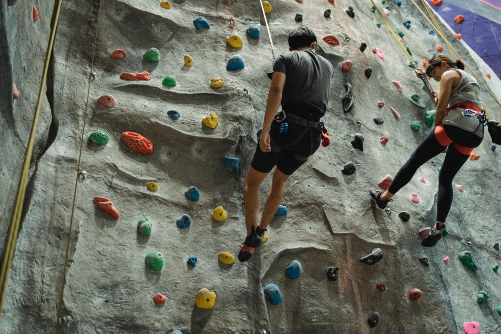 Two climbers on top-rope lines scaling a gray indoor climbing wall with colorful holds