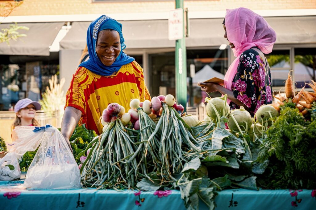 Two women talking across a table of spring onions and greens at an outdoor market