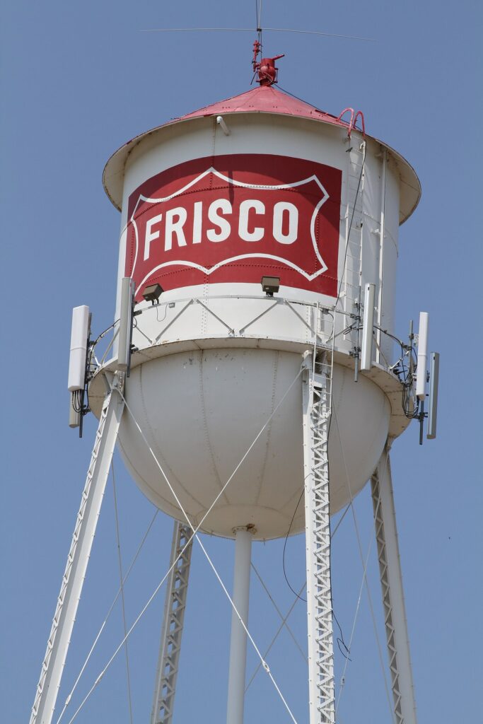 White downtown water tower with a red FRISCO logo against a clear blue sky