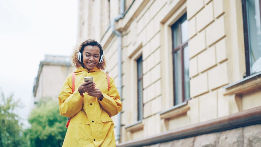 Woman in a yellow raincoat wearing headphones and looking at her smartphone outdoors