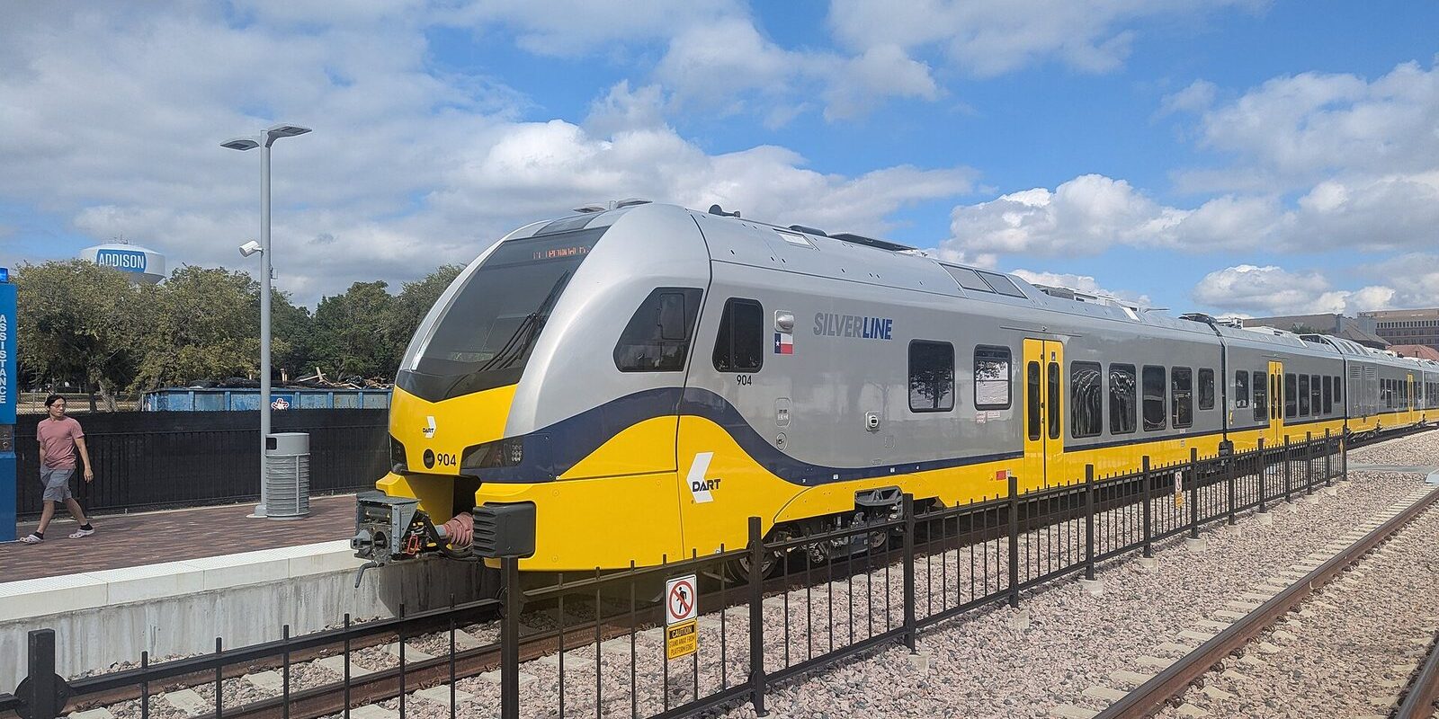 Yellow and silver DART Silver Line train stopped at a station platform under a partly cloudy sky